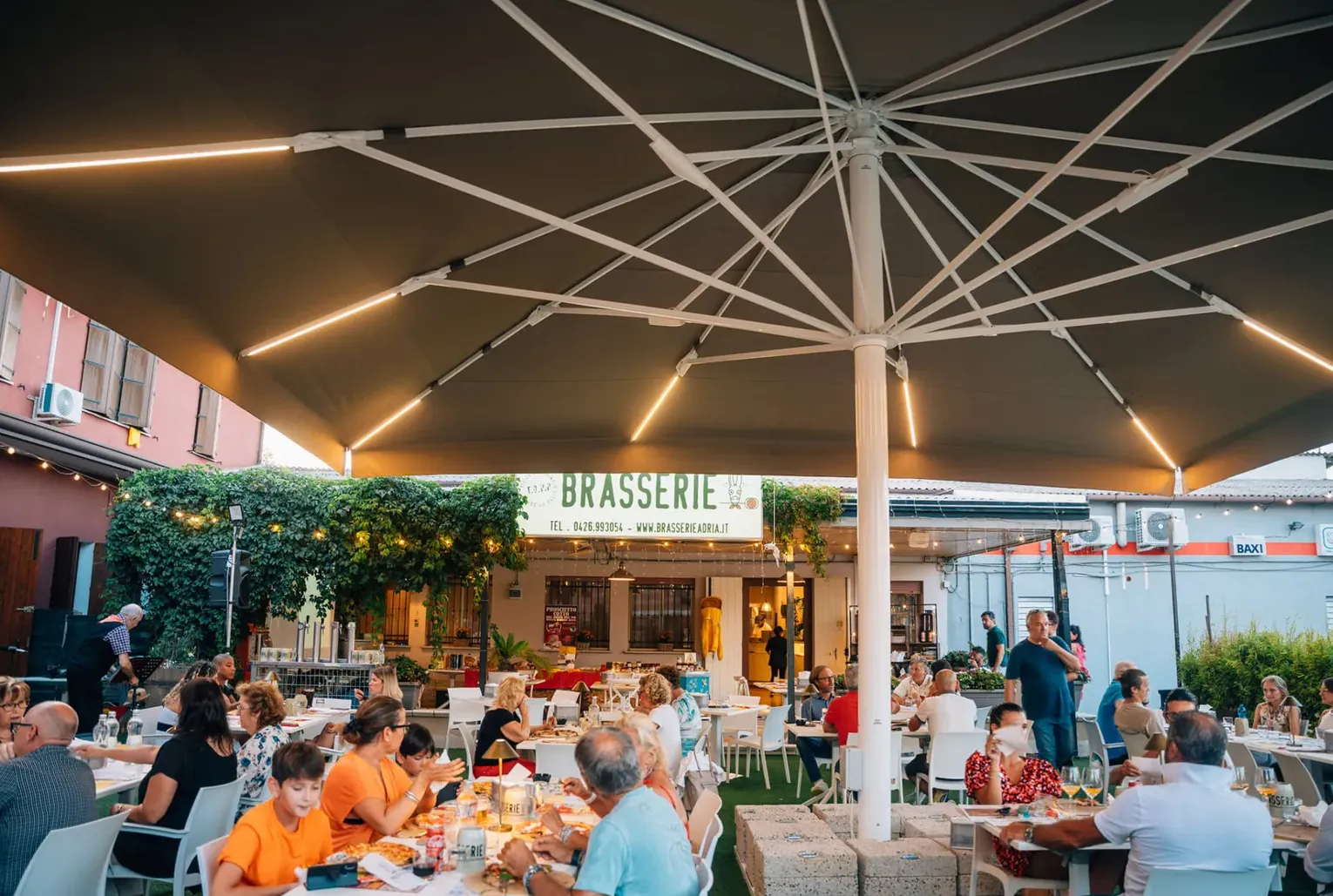 Il giardino della Brasserie pieno di gente sotto il grande ombrellone illuminato, con l'insegna sullo sfondo