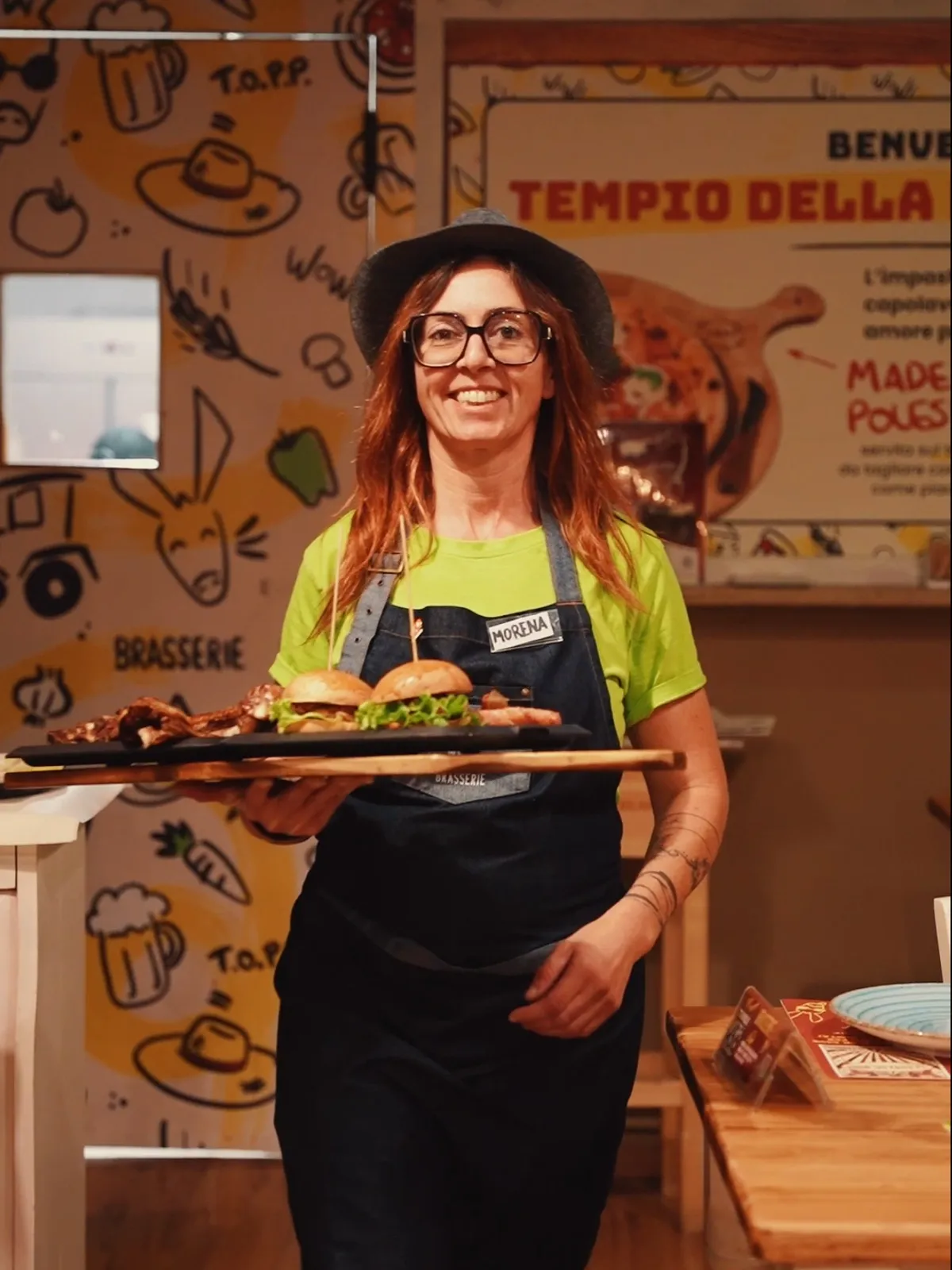 Una cameriera della Brasserie col cappello sorride e porta in sala il tagliere della Porcata con carne di maiale e burger